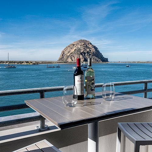 A table with two wine bottles and glasses on a balcony overlooking a body of water with boats and a large rock formation in the background.