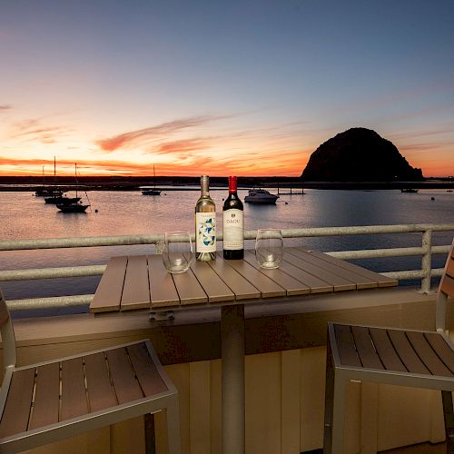 A sunset view over a bay with boats, a rock formation in the background, and a table with two wine bottles and glasses beside two chairs.