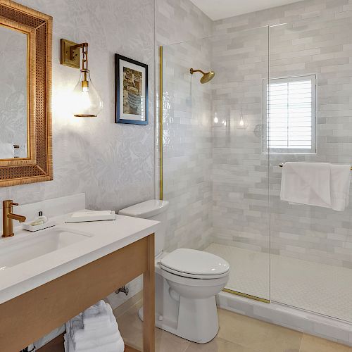 This image shows a modern bathroom with a shower, toilet, and sink. There's a large mirror, towel rack, and framed picture on the wall.