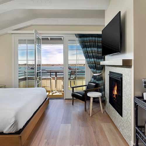 A modern hotel room features a bed, a fireplace, a TV, a small coffee station, and a balcony with a view of the water.