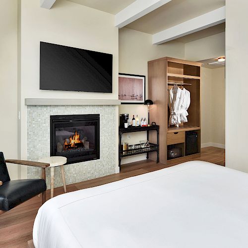 The image shows a modern hotel room with a bed, a wall-mounted TV above a fireplace, a chair, a side table, and an open wardrobe with robes.