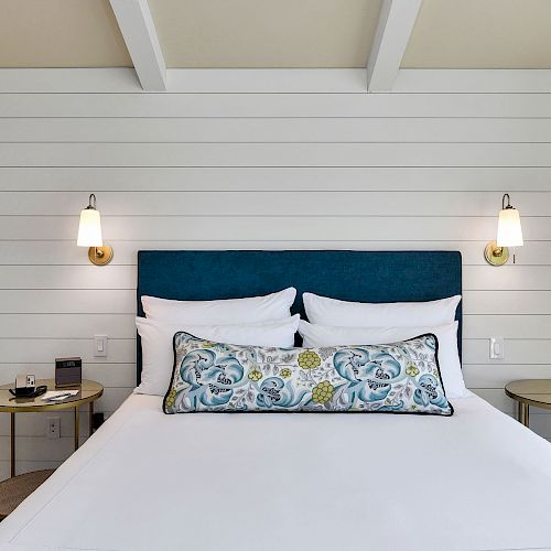 A cozy bedroom features a neatly made bed with blue and white bedding, two side tables, wall lamps, and a decorative painting on the wall.