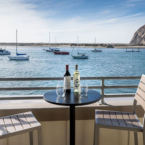 A table with two chairs overlooks the ocean, with bottles of wine and glasses on the table. Sailboats are in the water and a large rock formation.