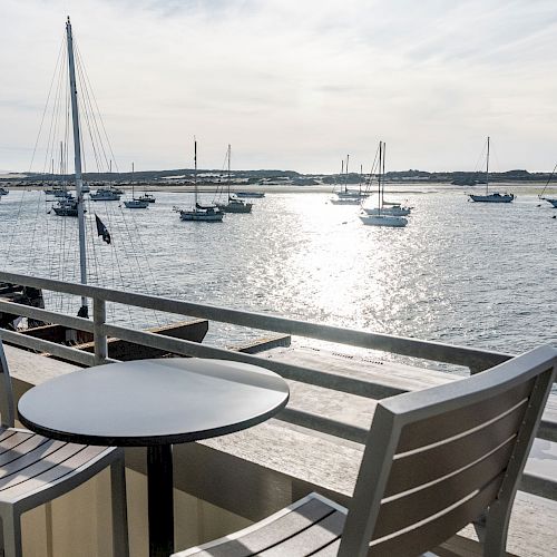 Balcony view with a round table and two chairs overlooking a calm sea with several sailboats under a cloudy sky in the distance.