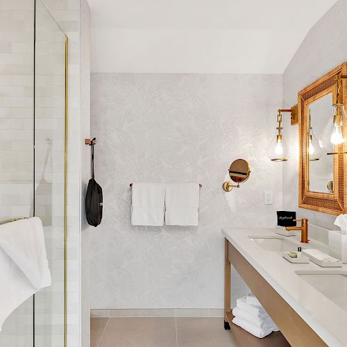 A modern bathroom features a glass shower, double sinks with golden faucets, framed mirrors, and wall-mounted lamps. Towels hang on the wall.