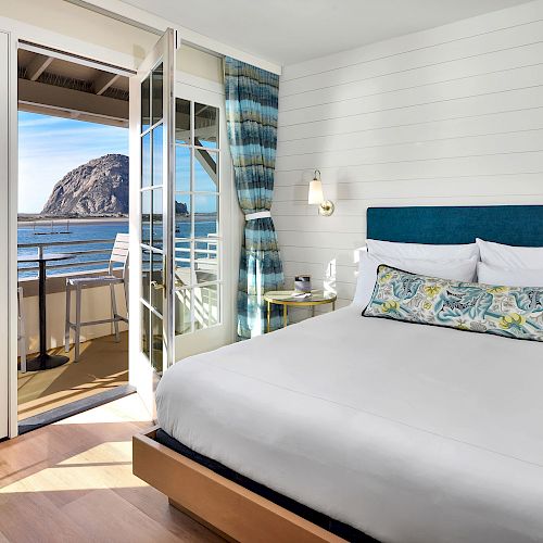 A cozy bedroom with a large bed, sliding glass doors leading to a balcony with chairs, and a scenic view of a mountain and water outside.