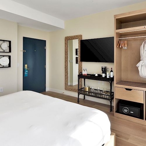 A modern hotel room with a neatly made bed, TV, robes, mini-fridge, safe, mirror, and a glimpse of a bathroom on the left.