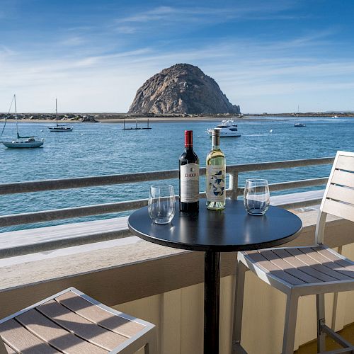 A scenic view of a waterfront with a table, two chairs, two wine bottles, and glasses. A large rock formation and boats are visible in the distance.