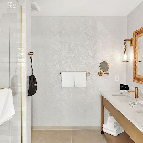 A clean, modern bathroom with a glass shower, vanity with double sinks, golden fixtures, framed mirrors, and neatly folded towels.