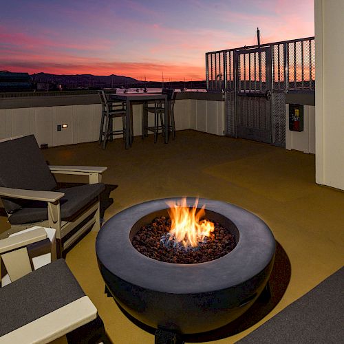 An outdoor patio with a fire pit, surrounded by chairs and tables, overlooking a scenic sunset.