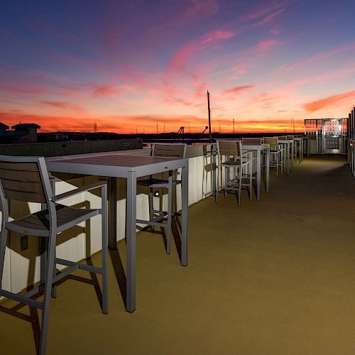 The image shows a rooftop terrace with tables and chairs, set against a stunning sunset sky with vibrant colors, creating a peaceful atmosphere.