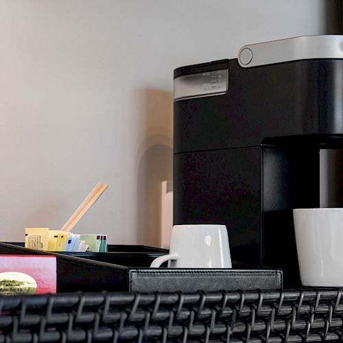 The image shows a coffee station with a Keurig coffee machine, two white mugs, bottled water, stir sticks, and a small tray with condiments.