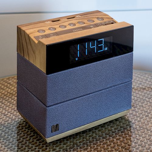 The image shows a digital alarm clock with a wooden and fabric design, displaying the time 11:43 on a reflective surface.