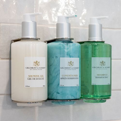 Three bottles of toiletries mounted on a tiled wall: shower gel, conditioner, and shampoo, with pumps on top, labeled 