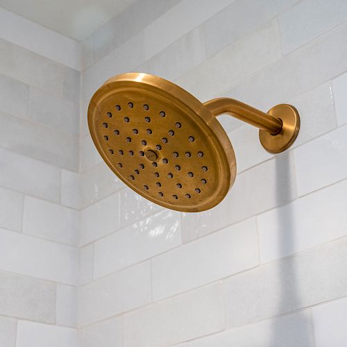 The image shows a gold-colored, round showerhead mounted on a wall with white tiles in a bathroom.