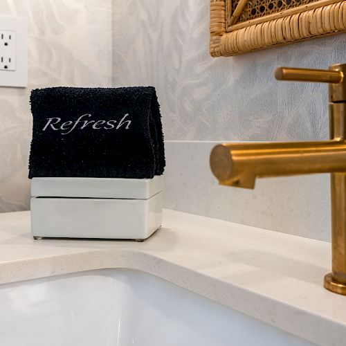 The image shows a bathroom sink area with a gold-colored faucet, a mirror with a woven frame, and a black towel labeled 