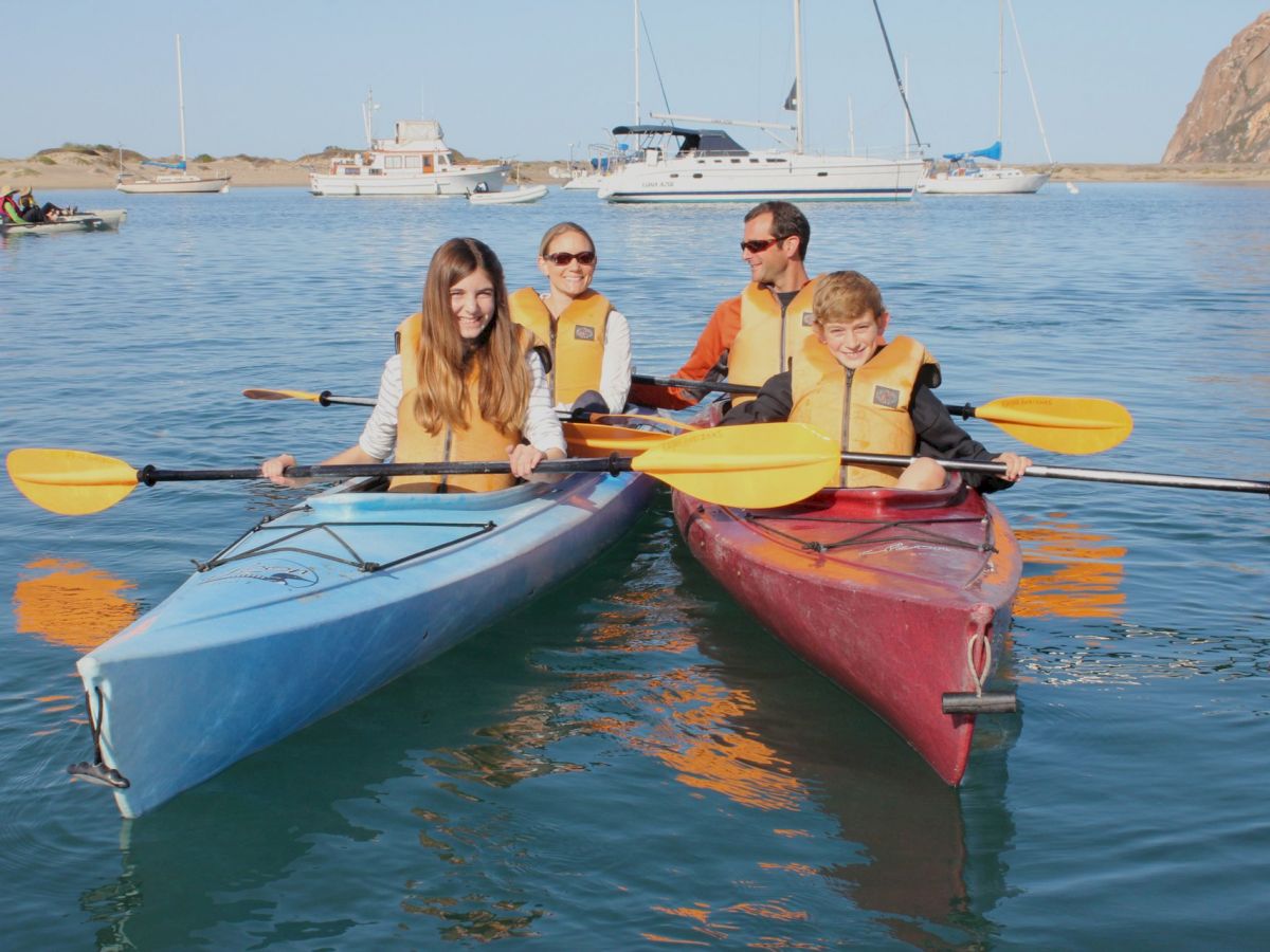 Four people in life jackets are kayaking in two kayaks on a calm water body with boats and a rocky shoreline in the background.