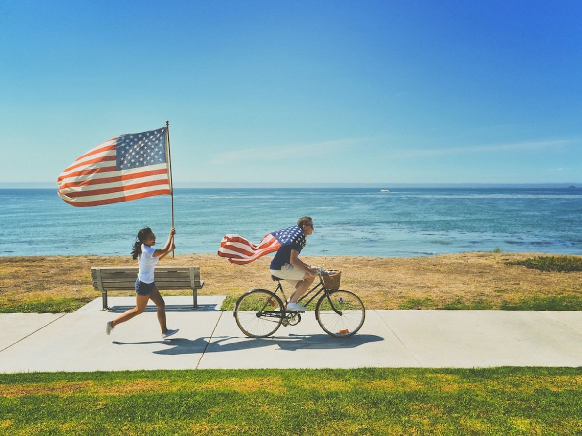 Two people are enjoying a sunny day by the ocean, one riding a bicycle and the other running, both holding American flags.