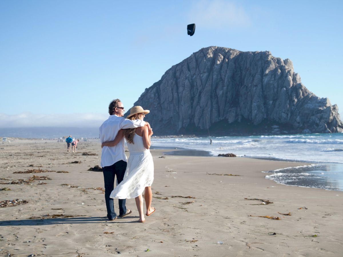 A couple walks arm in arm on a sandy beach with a large rocky mountain in the background, with clear skies and gentle waves ending the sentence.