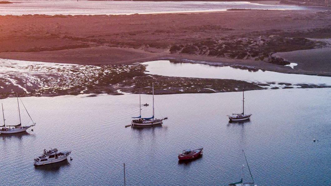 A serene sunset over a coastal lagoon, with several boats anchored on calm water and a sandy shore in the background.