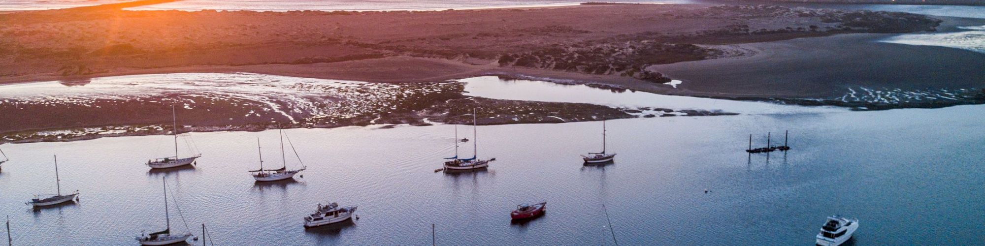 A serene sunset over a coastal lagoon, with several boats anchored on calm water and a sandy shore in the background.