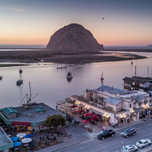 Coastal scene with boats, buildings, and a large rock formation at sunset. A street lined with cars and businesses is in the foreground.