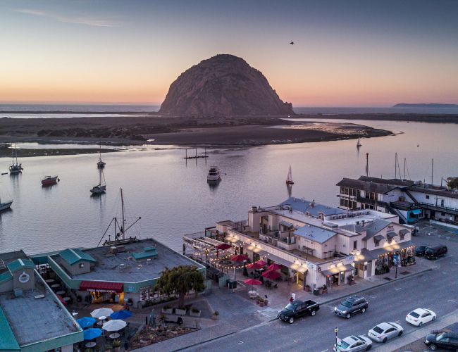 Coastal scene with boats, buildings, and a large rock formation at sunset. A street lined with cars and businesses is in the foreground.