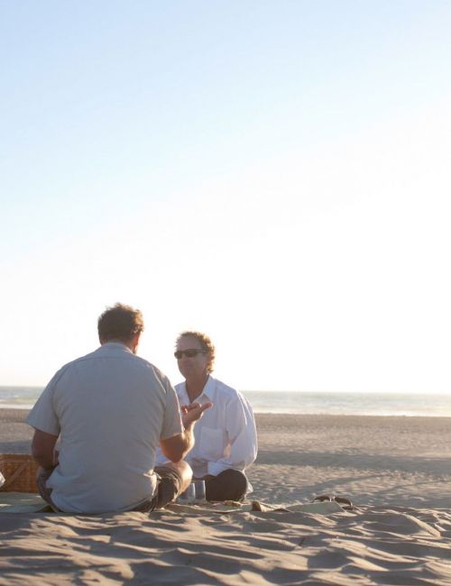 Three people sitting on a sandy beach, with the ocean in the background and a few distant figures walking along the shore.