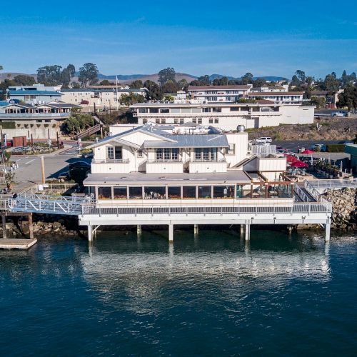 An oceanside building built on stilts over water, with surrounding coastal town and greenery visible in the background.