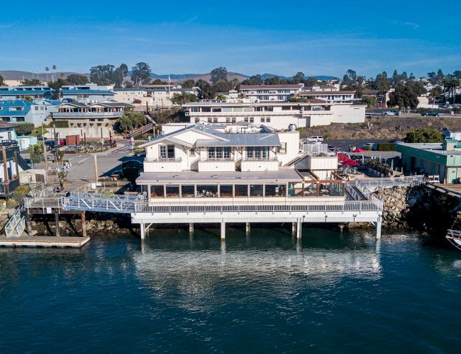 An oceanside building built on stilts over water, with surrounding coastal town and greenery visible in the background.
