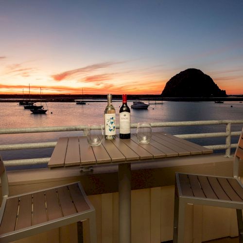 A sunset view with two chairs and a table holding wine bottles and glasses, overlooking a bay with boats and a large rock formation.