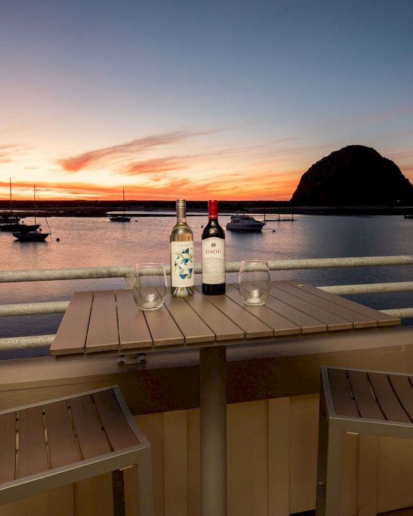 A sunset view with two chairs and a table holding wine bottles and glasses, overlooking a bay with boats and a large rock formation.