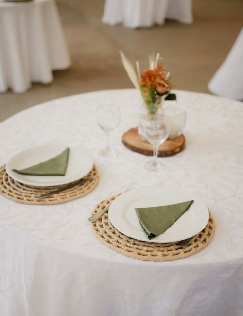 A round table with a white cloth, two place settings with green napkins, wine glasses, and a small floral centerpiece is shown.