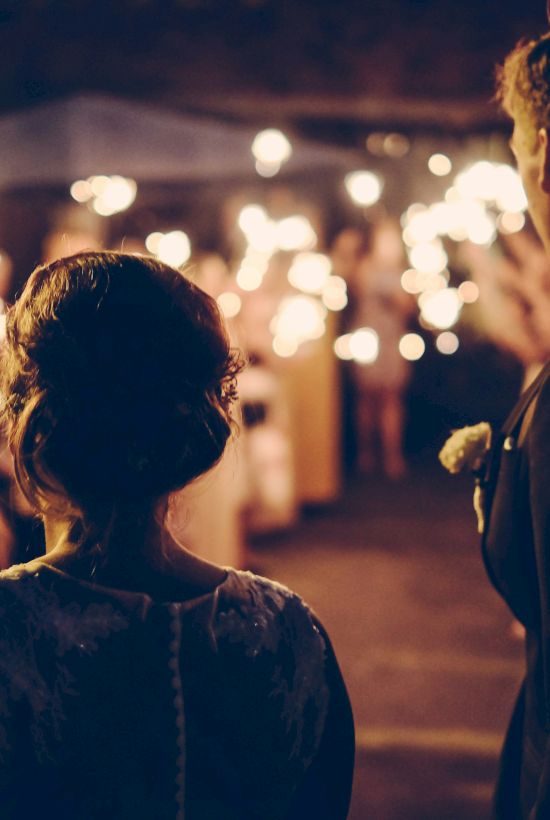 A couple stands together with guests holding sparklers at a nighttime celebration, possibly a wedding or festive event, creating a warm, lively atmosphere.