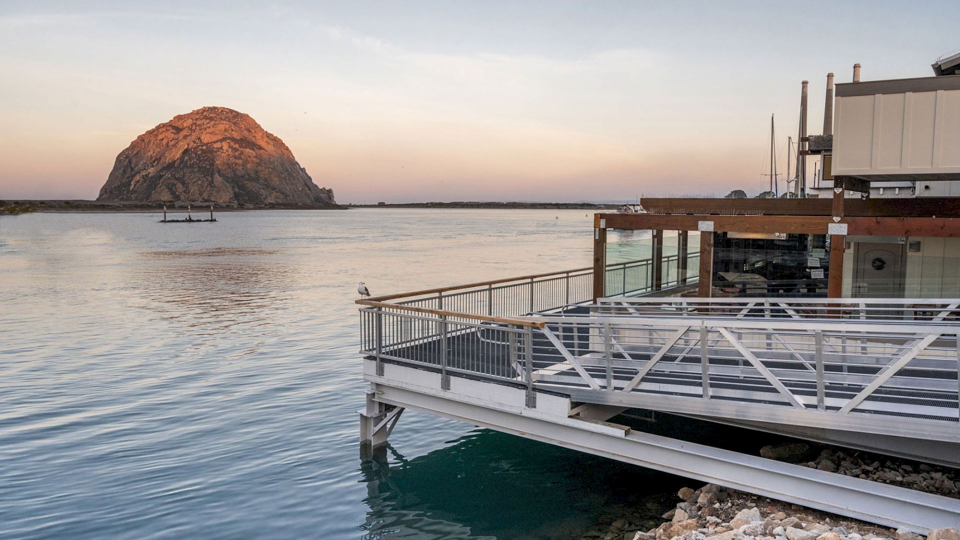 A peaceful waterfront view with a large rock formation in the distance and a building with a deck by the water&rsquo;s edge at sunset.