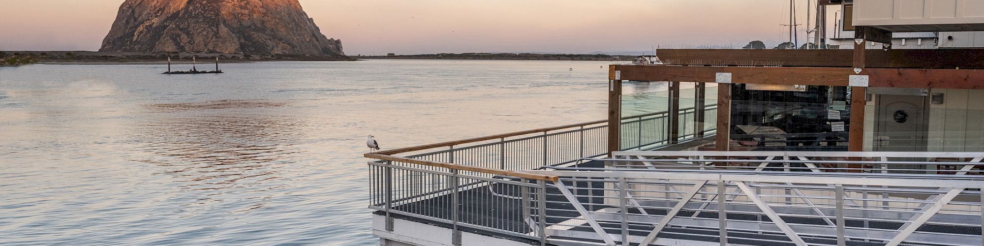 A peaceful waterfront view with a large rock formation in the distance and a building with a deck by the water’s edge at sunset.