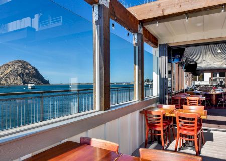A scenic view of a coastal restaurant patio with wooden furniture, overlooking the ocean and a large rock formation in the background.