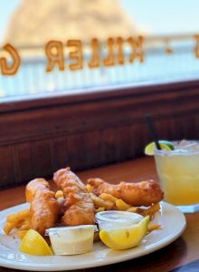 The image shows a plate of fish and chips with lemon wedges and dipping sauces, accompanied by a drink with lime garnish on a wooden table.