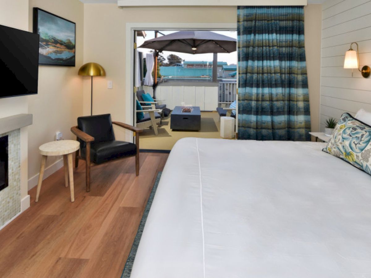 The image shows a cozy bedroom with a large bed, a chair, a TV, and an open sliding door leading to a patio with outdoor seating.