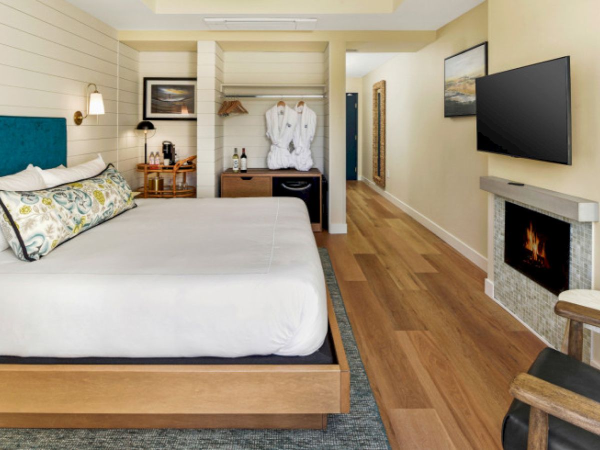 A cozy hotel room with a large bed, TV, fireplace, desk, and wardrobe, featuring wooden flooring and warm decor.