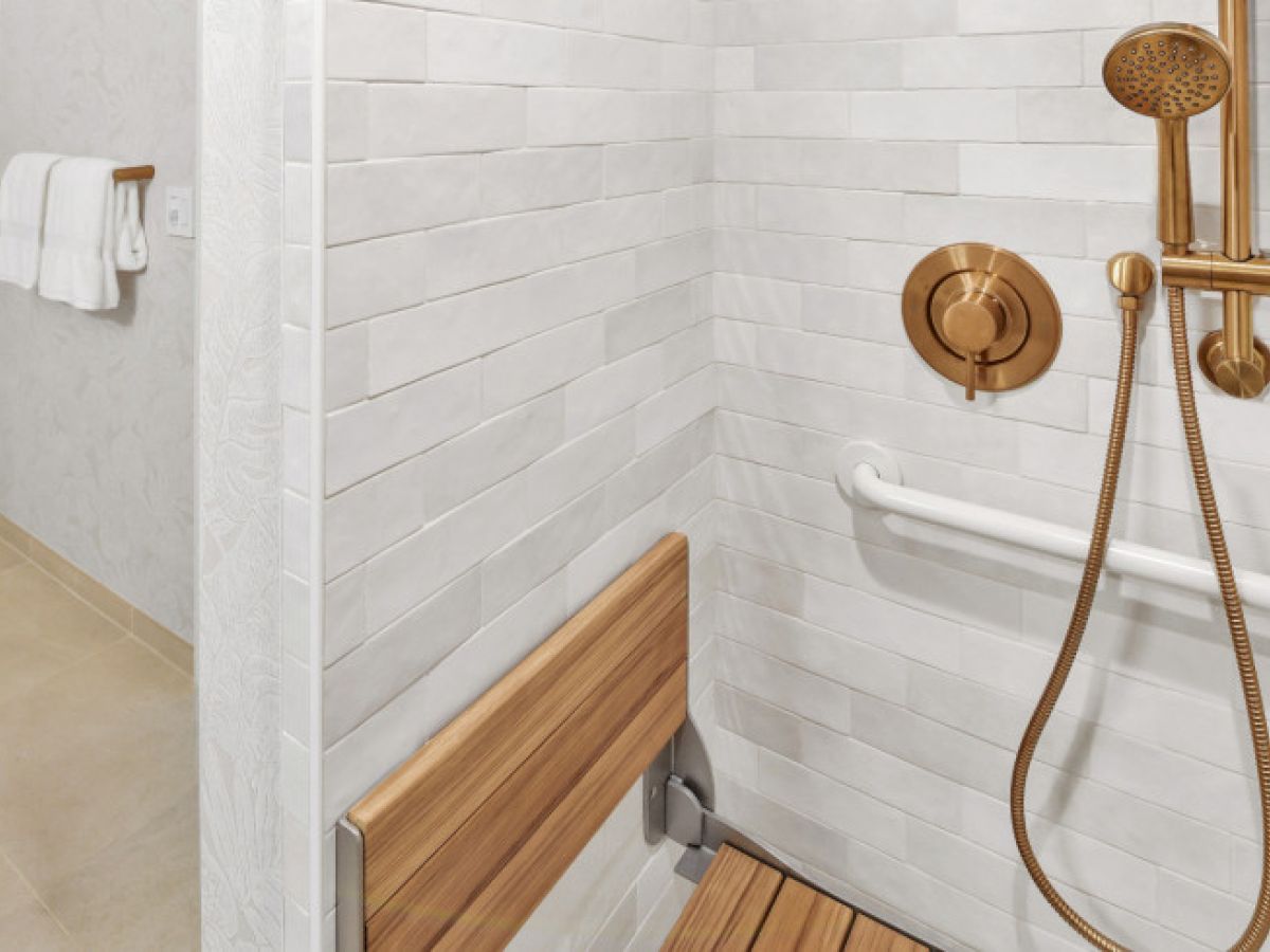 The image shows a bathroom with a shower area, featuring a wooden seat, gold-colored shower fixtures, and a towel rack nearby.