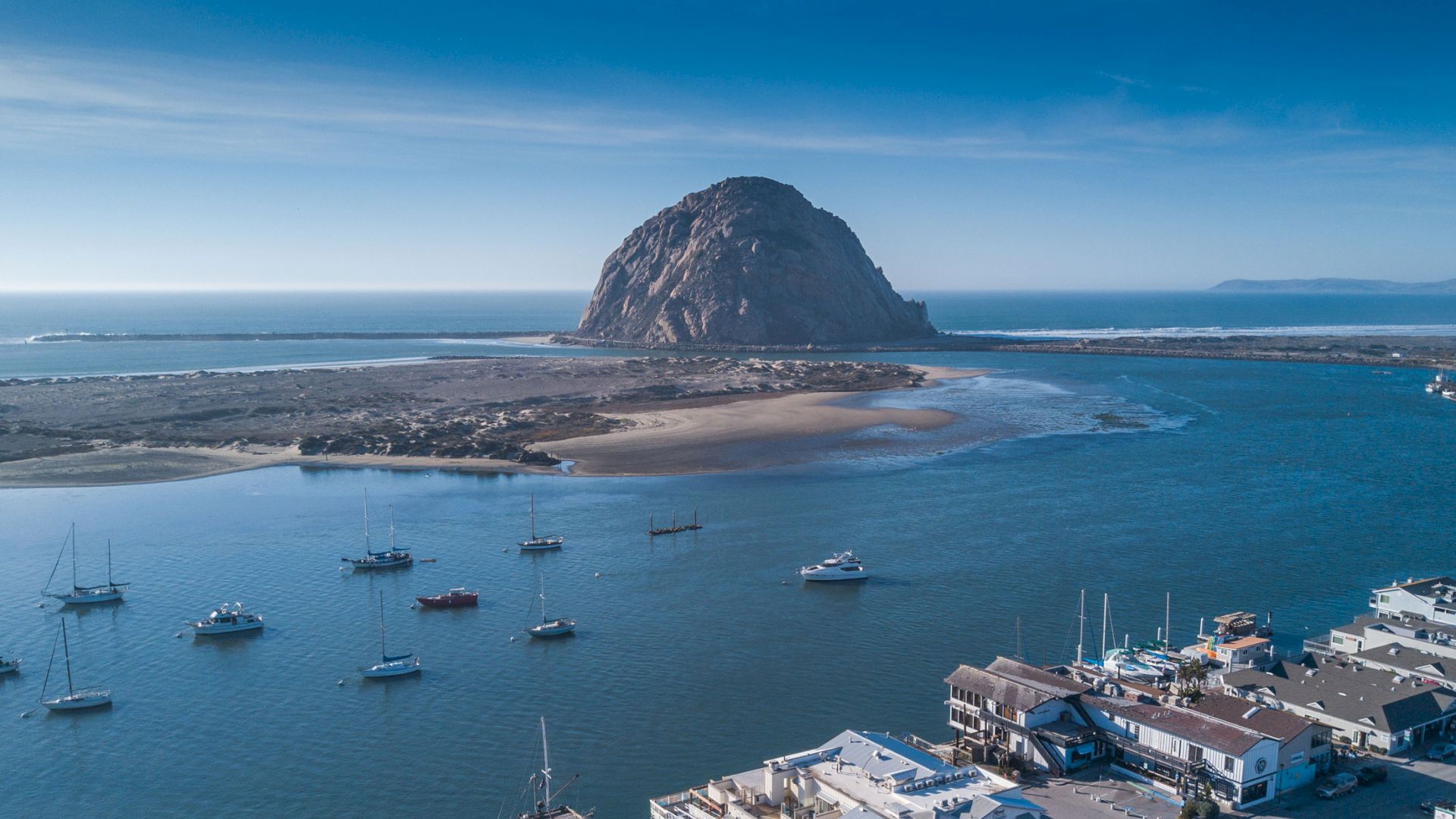 Colorful coastal town with a rocky island outcrop, boats in the harbor, and whitewashed buildings by the shore under a clear sky.