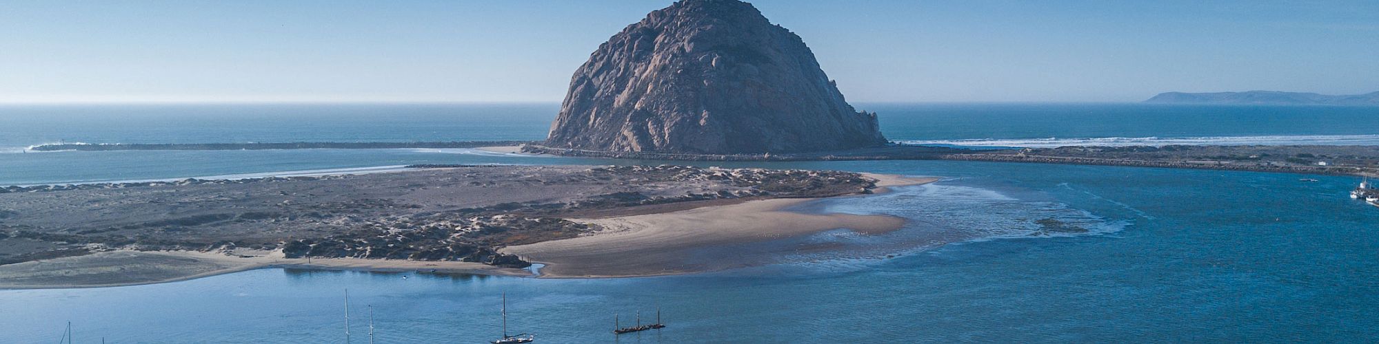 Colorful coastal town with a rocky island outcrop, boats in the harbor, and whitewashed buildings by the shore under a clear sky.