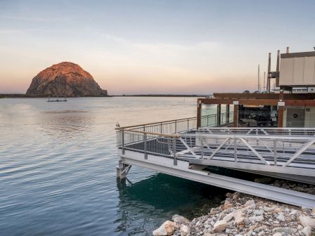 A calm waterfront scene at sunset with a rocky shore, a wooden dock extending over clear water, and a rocky island in the distance.
