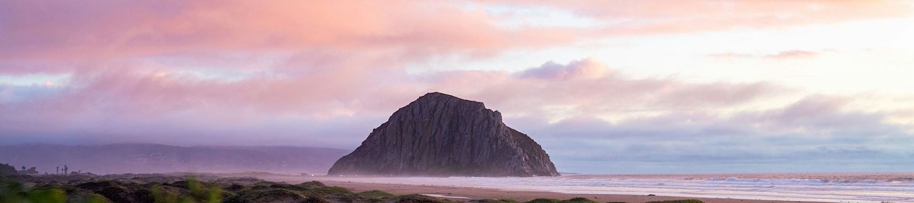 A serene beach at sunset with a lone rock outcrop rising from sand under pink-purple clouds.