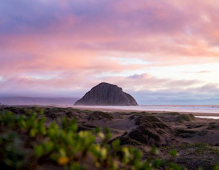 A serene beach at sunset with a lone rock outcrop rising from sand under pink-purple clouds.