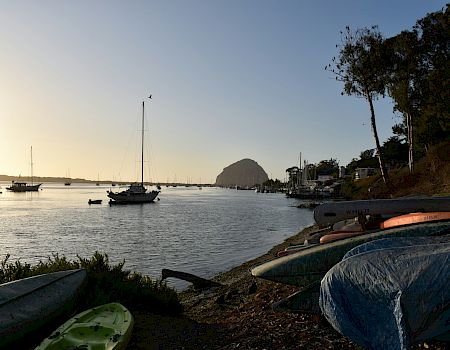 Sailboats anchored on a calm bay at sunset, with small boats/kayaks on the shore and a hill in the distance.