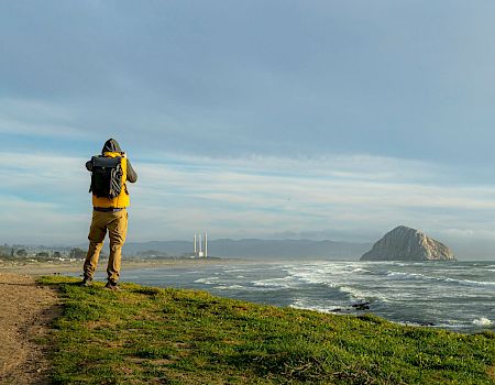 A hiker in yellow/black gear with a large backpack stands by the ocean, looking toward waves and distant hills.