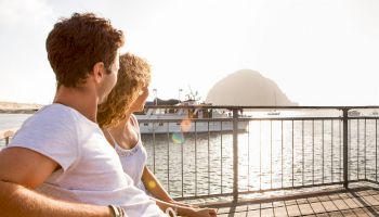 A couple sitting on a bench by the water, looking at a docked boat with a large rock formation in the background, under a sunny sky.