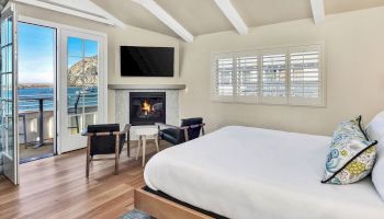 A cozy bedroom features a bed, two chairs, a fireplace, TV, and a balcony with ocean views, all under a vaulted ceiling with white beams.